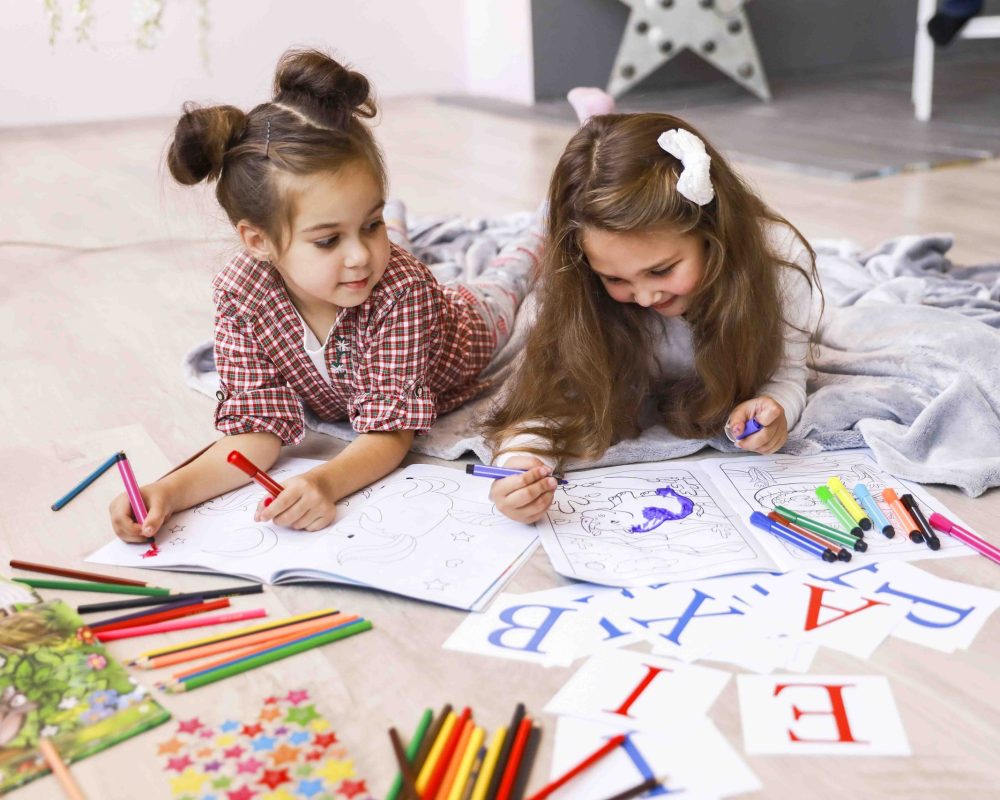 Two tiny happy girls which are drawing in the coloring book laying on the floor on the blanket and learning letters